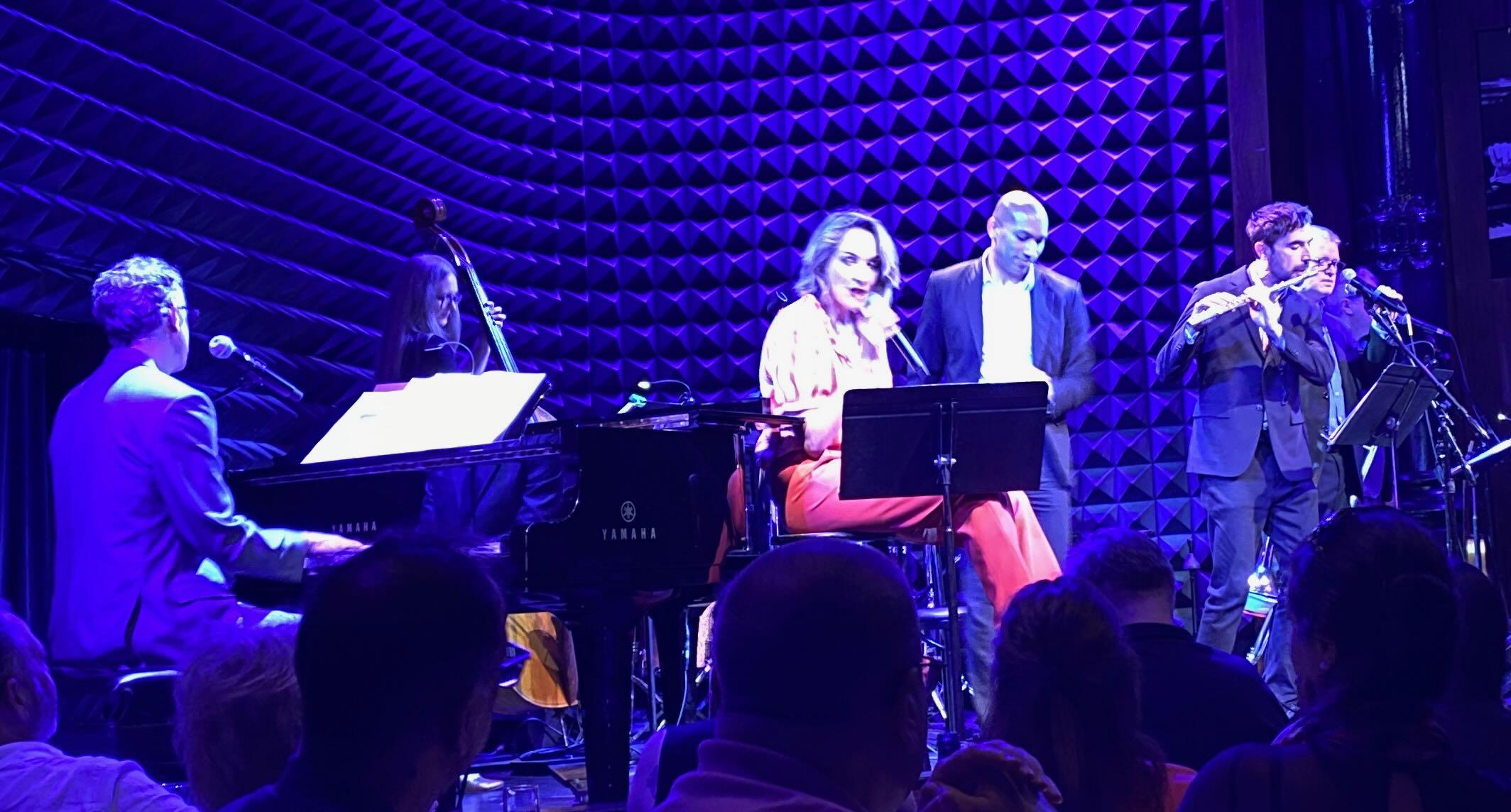 Photo shows a group of musicians performing in a darkened club.  The blonde female lead singer sits on a stool singing into a microphone and looking into the crowd.  To left a piano player is seen from behind. Beyond the piano is a young female bass player. To the right of the singer is a tap dancer and a flutist. Obscured are two trombone players adn the drummer.