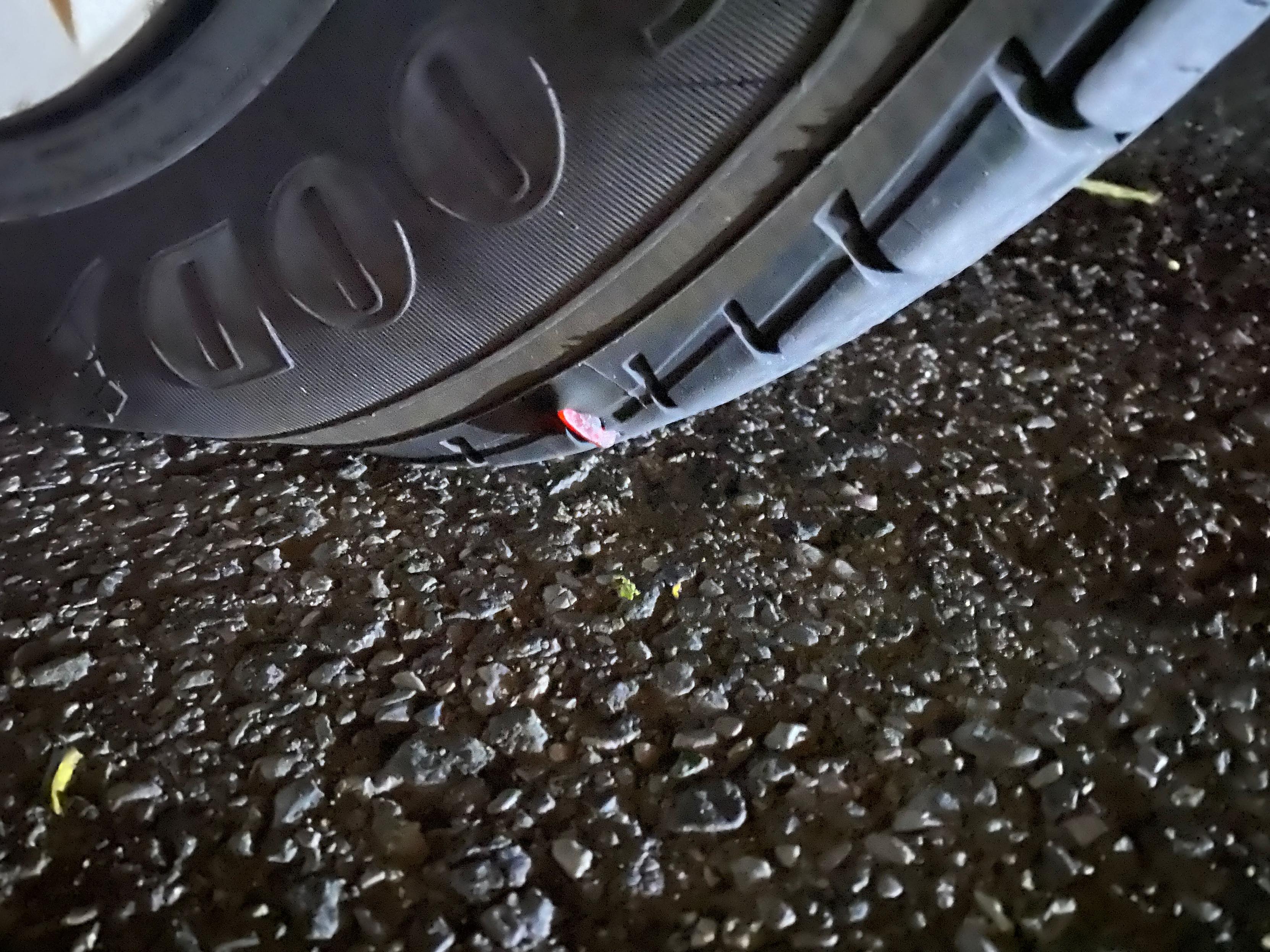 Photo is a close up of a partially flat tire on a darkened asphalt street.  A sharp piece of red plastic is seen stuck at the edge of the treat, too close to the side wall. unfortunately, this won't be able to be fixed. Going to be big $$ for new tires (can't just replace 1).  Likely picked this up in the parking garage, as the TPMS warning came on moments after leaving. It could be worse though,  it could have been on the way home, in the dark with no where to pull off the road.